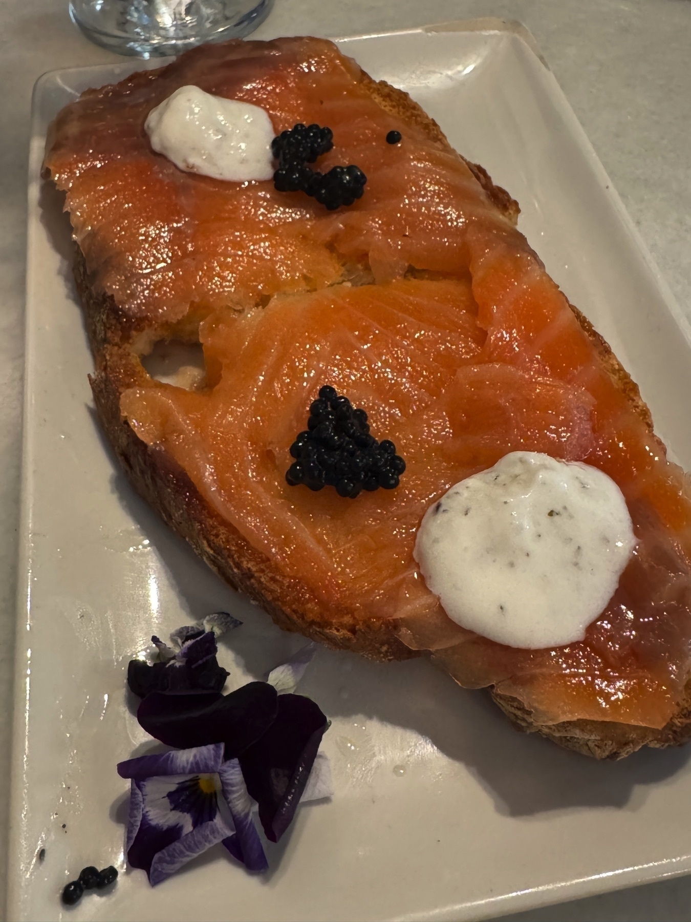 Tosta de salmón con tzatziki, featuring a large slice of toasted bread topped with silky smoked salmon, creamy dollops of tzatziki sauce, and spoonfuls of black caviar. Garnished with purple edible flowers, the dish offers a luxurious combination of color, texture, and rich flavors.