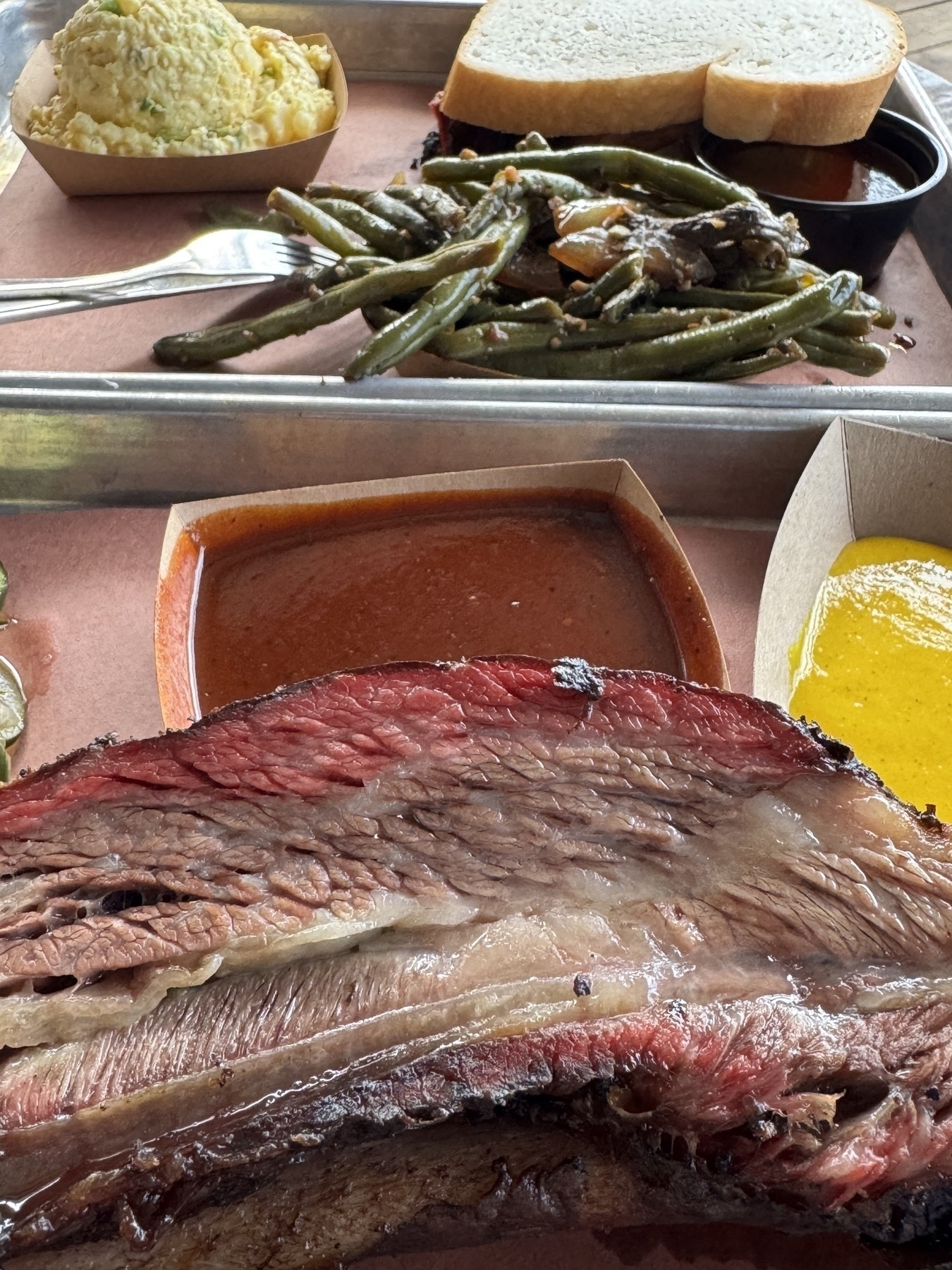 Close-up of a barbecue tray featuring a large smoked beef rib with a well-defined smoke ring, accompanied by green beans, white bread slices, potato salad, yellow mustard-based sauce, and a reddish-brown barbecue sauce.