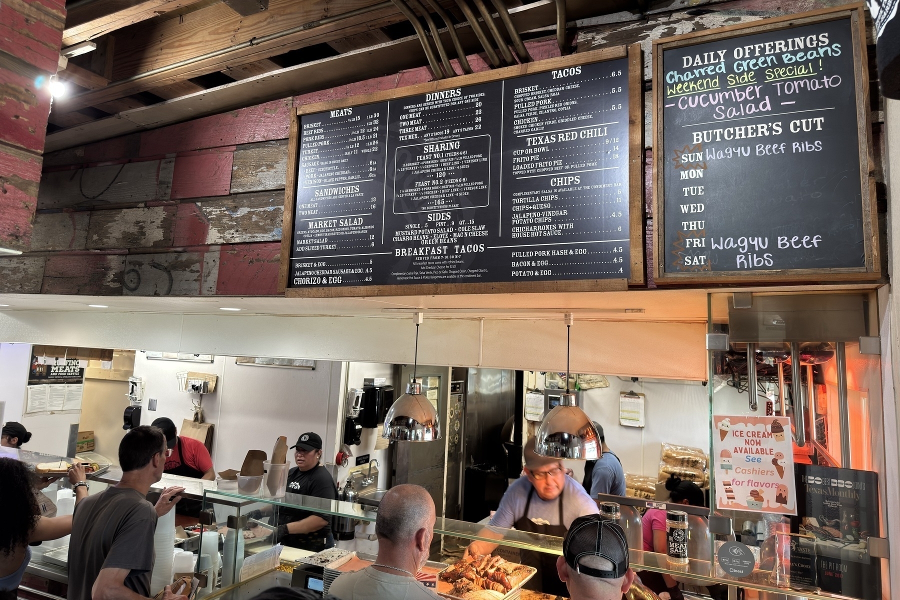 Interior of The Pit Room showing customers in line at the counter as staff serve barbecue behind glass. Overhead, two large menu boards display offerings like brisket, beef ribs, tacos, and sides, with a chalkboard listing “Wagyu Beef Ribs” as the weekend special.