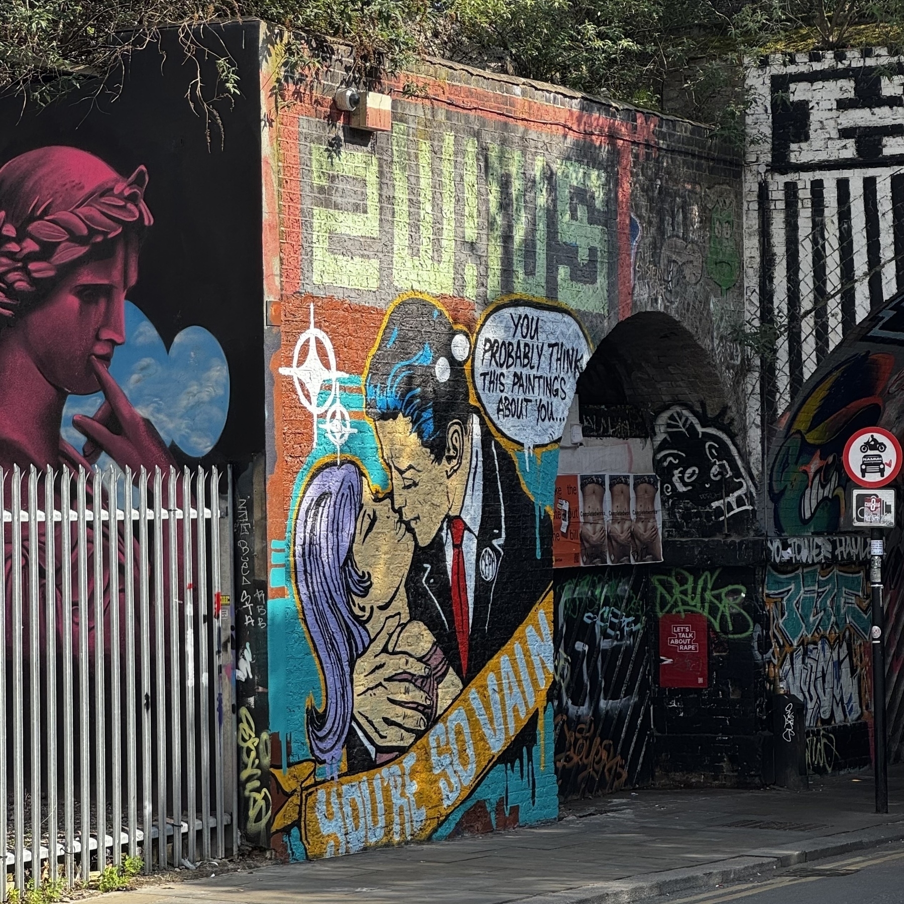 Graffiti mural shows a couple kissing with comic-style text reading “You probably think this painting’s about you” and “You’re so vain,” surrounded by layered street art and tags.