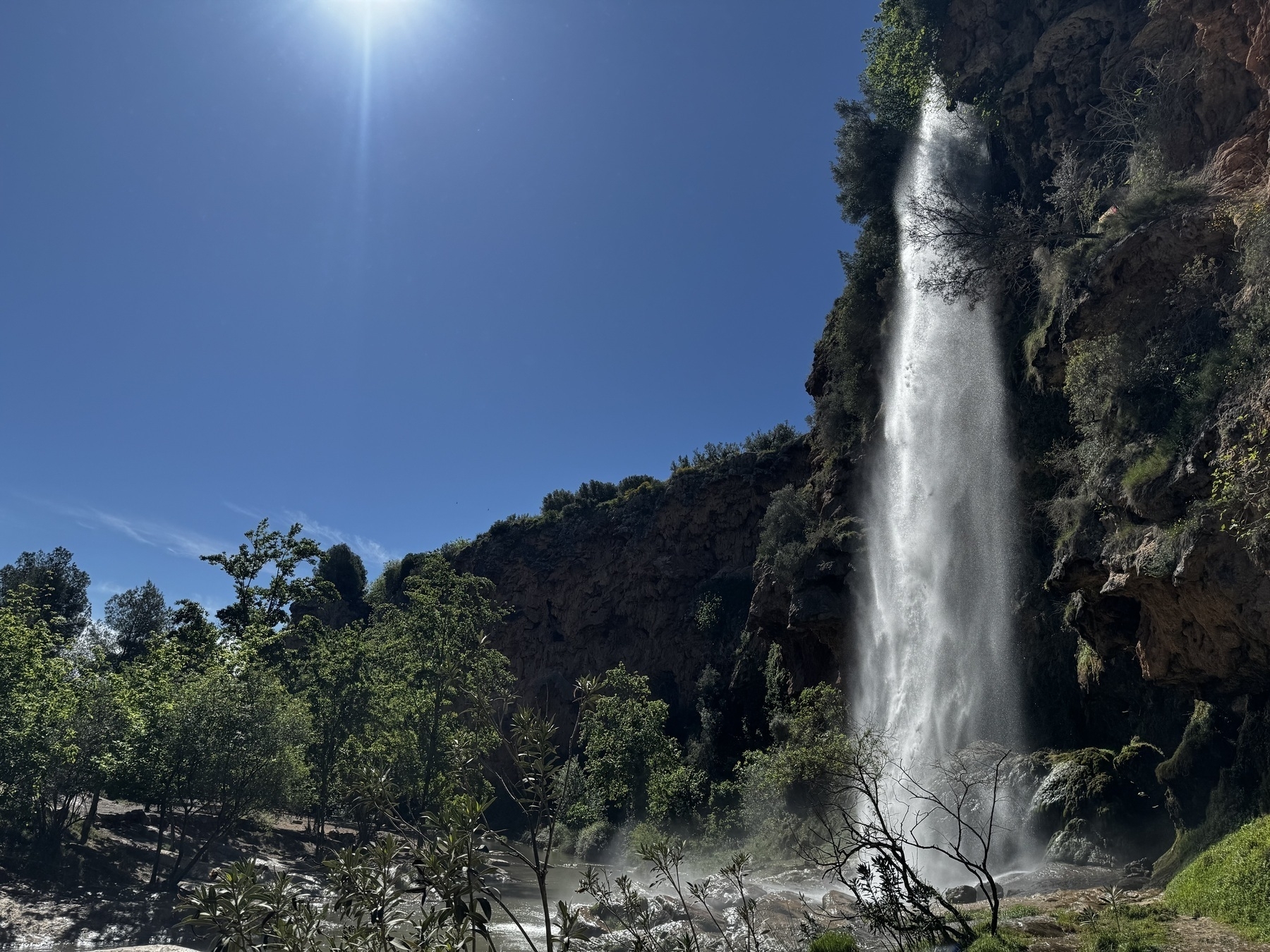 A tall, narrow waterfall plunges from a cliff surrounded by lush greenery under a bright blue sky with the sun overhead, creating mist at the bottom.