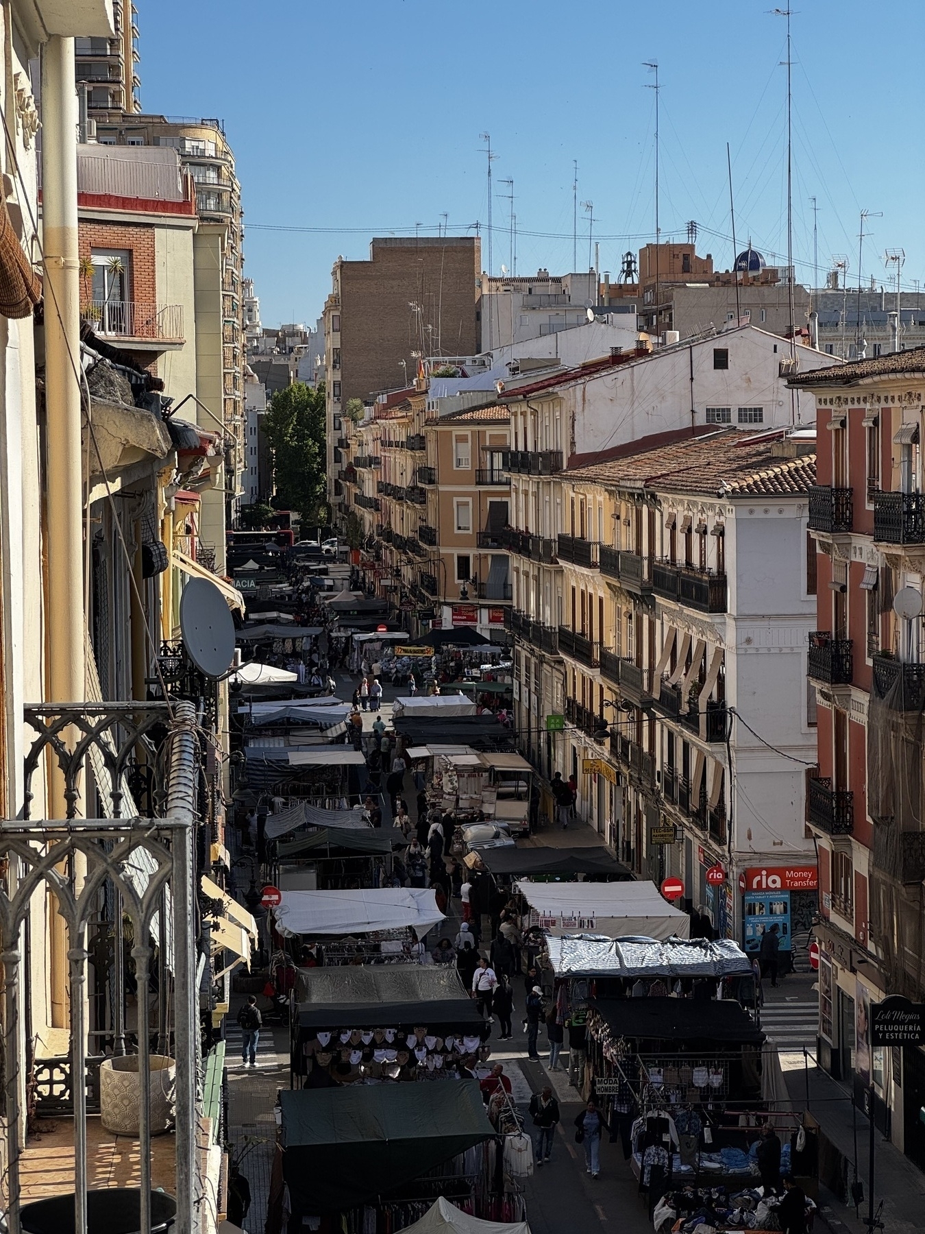 View from a balcony overlooking a lively street market in a residential neighborhood. The narrow road is lined with vendor stalls shaded by canopies, offering clothing and various goods. Pedestrians fill the street, weaving between the tightly packed stalls. Mid-rise apartment buildings with balconies and shuttered windows frame the scene under a clear blue sky.