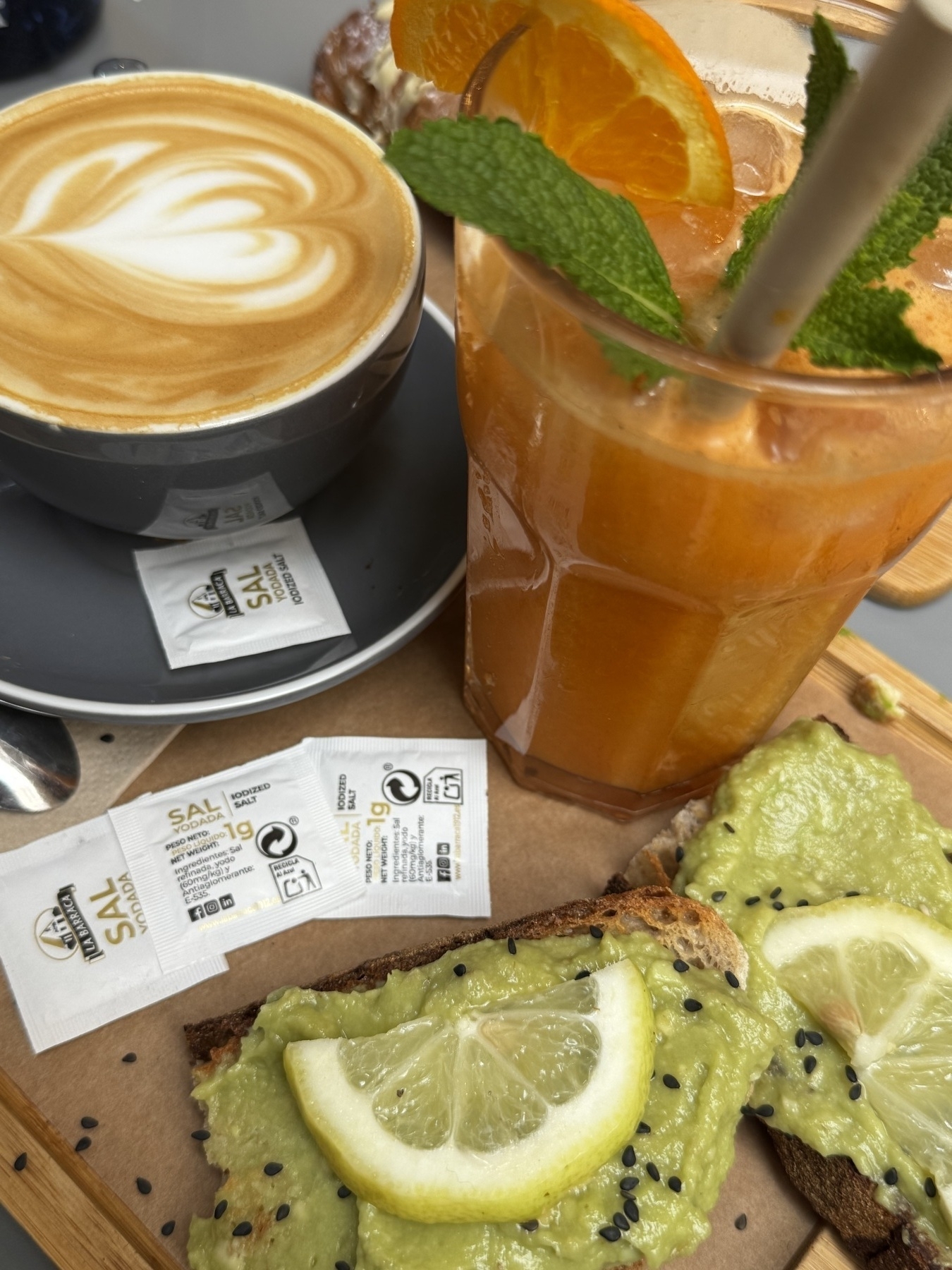 Close-up of a breakfast spread on a café table, featuring a cappuccino in a gray cup with heart-shaped foam art, a tall glass of orange juice garnished with mint sprigs and dried orange slices, and two slices of dark toast topped with mashed avocado, black sesame seeds, and lemon wedges. Individual packets of iodized salt rest on the wooden tray.
