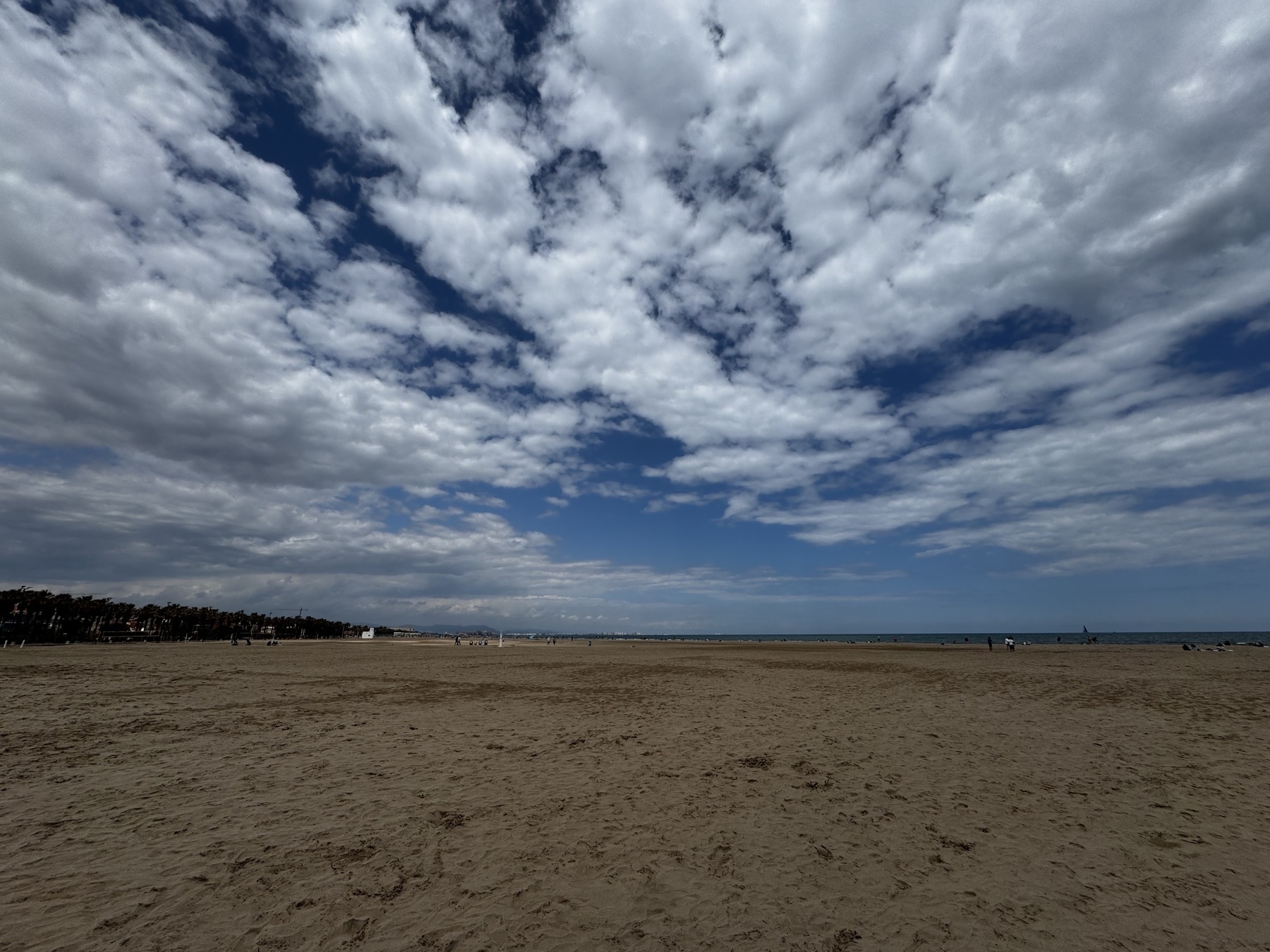 Wide view of a vast sandy beach beneath a dramatic, cloud-filled sky. The clouds stretch in textured layers across the frame, revealing patches of blue sky. A calm, flat sea meets the sand in the distance, where a few scattered beachgoers are visible. The setting feels spacious and open.