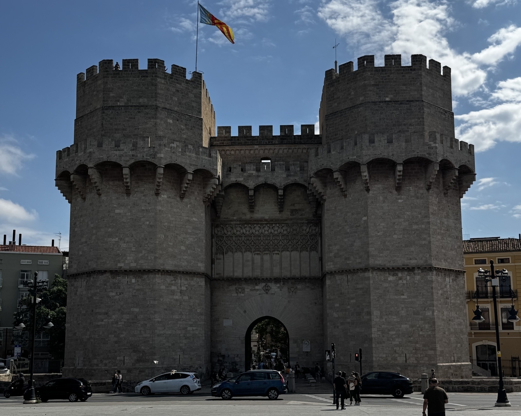 The Torres de Serranos, a medieval city gate with two large stone towers joined by a central archway and topped with battlements. The Gothic structure features intricate carved details above the arched passageway. A regional flag flutters atop one tower. People walk and cars pass in front of the landmark, partially shaded by afternoon light.