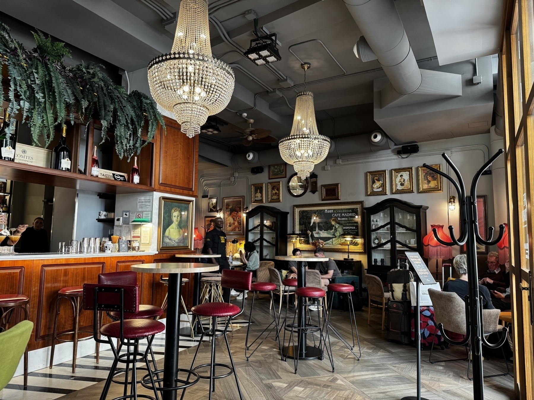Interior of Café Madrid, a stylish bar and café with classic European flair. Two grand chandeliers hang from the ceiling, illuminating a mix of red velvet bar stools, marble-top tables, and vintage framed artwork on the walls. The wooden bar is stocked with wine and spirits, and patrons are seated in relaxed conversation around the room.