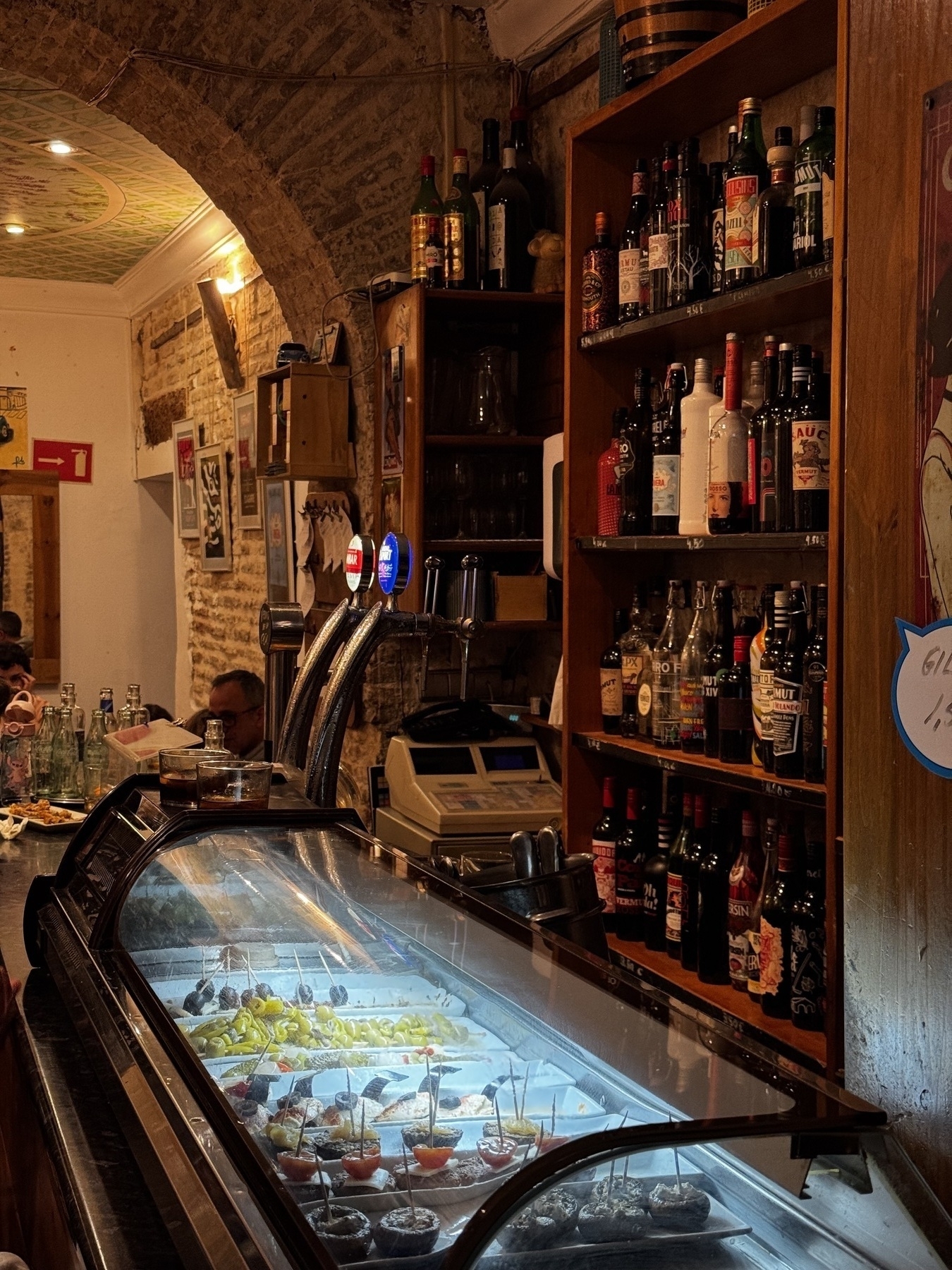 Inside a traditional vermouth bar with arched brick ceilings and rustic décor. A curved glass display case holds an array of pintxos, including skewered olives, peppers, and cured meats. Behind the counter, shelves are filled with bottles of liquor and vermouth. Warm lighting and old-world charm set a cozy, intimate atmosphere as patrons enjoy drinks.
