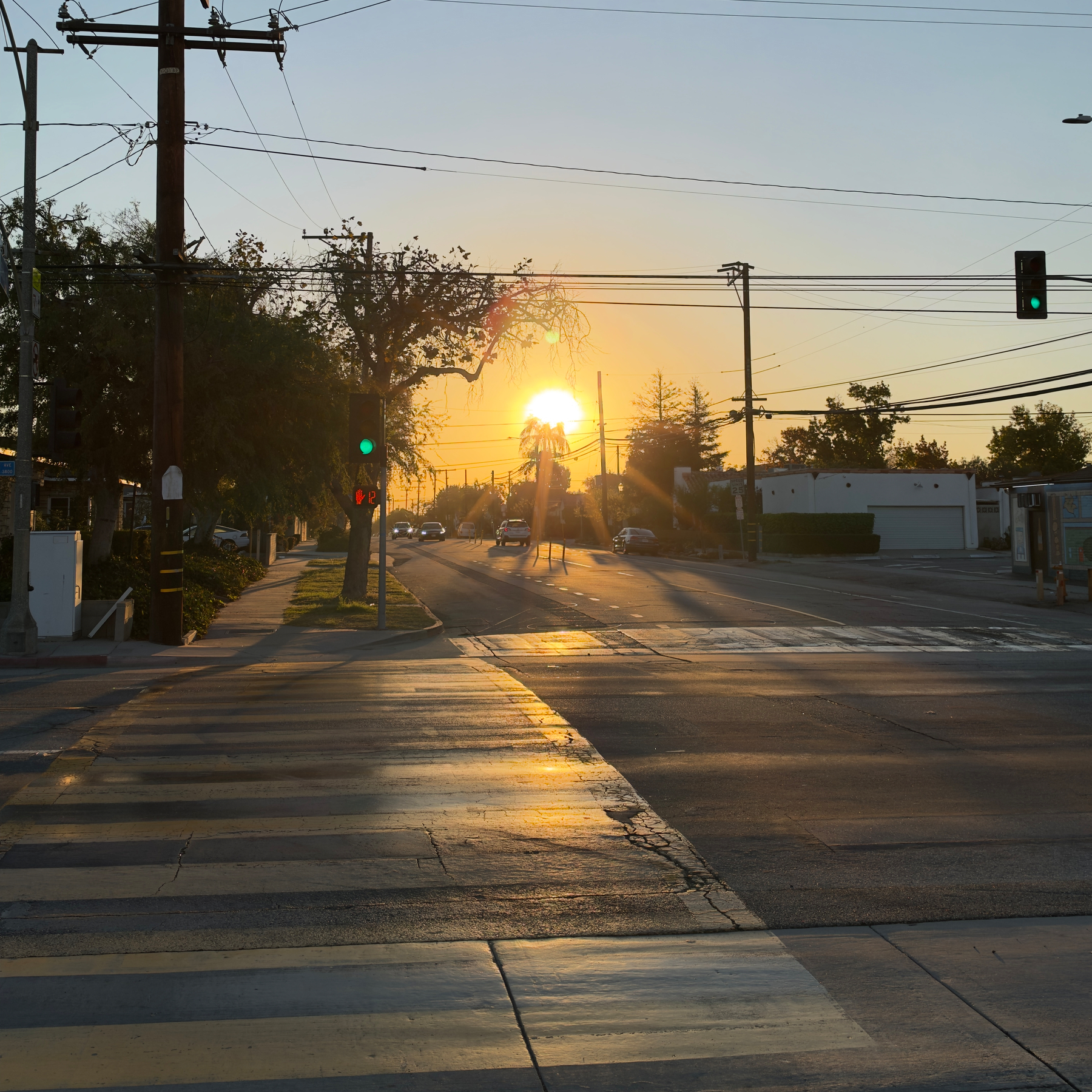 A street intersection with a pedestrian crosswalk during sunrise, showing green traffic lights and some vehicles in the distance.